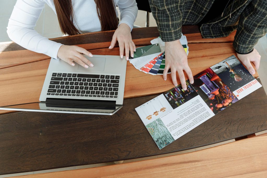 Two colleagues working on a design project using a laptop and color guides at a wooden table.
